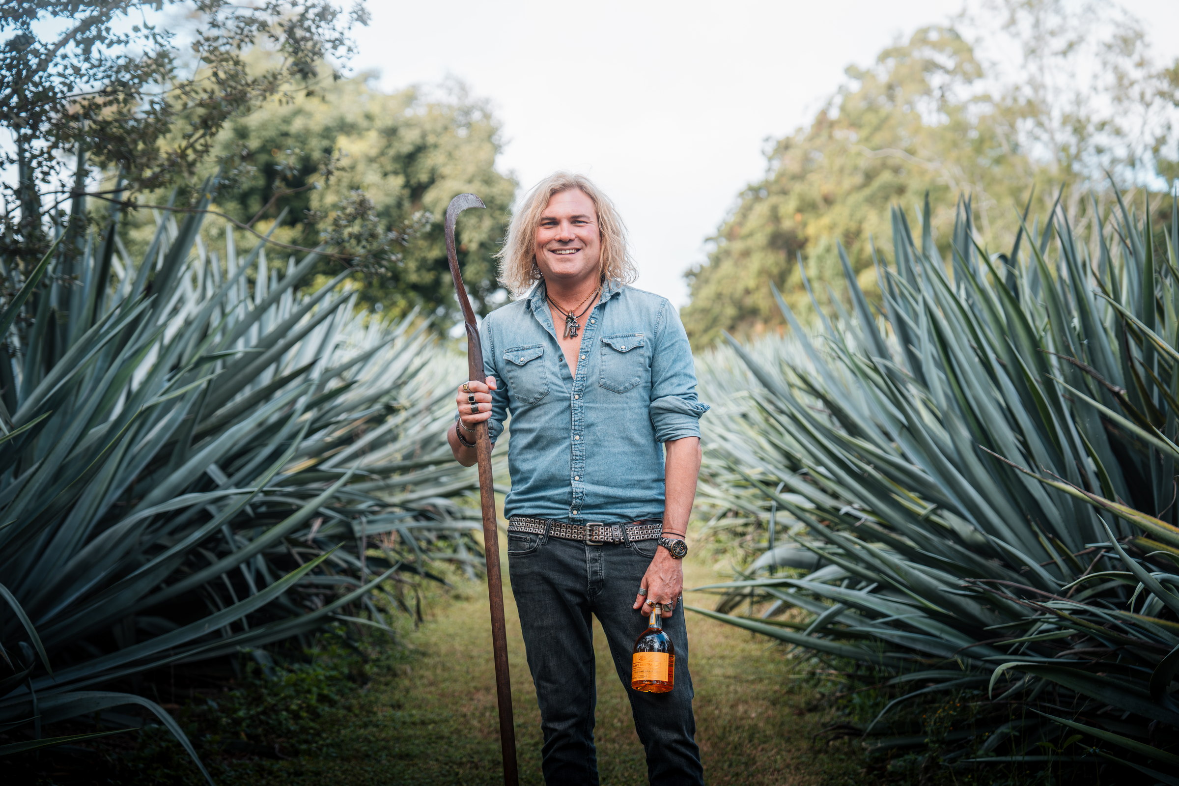 The grower standing between rows of blue agave holding a coa and a bottle of Agave Australis