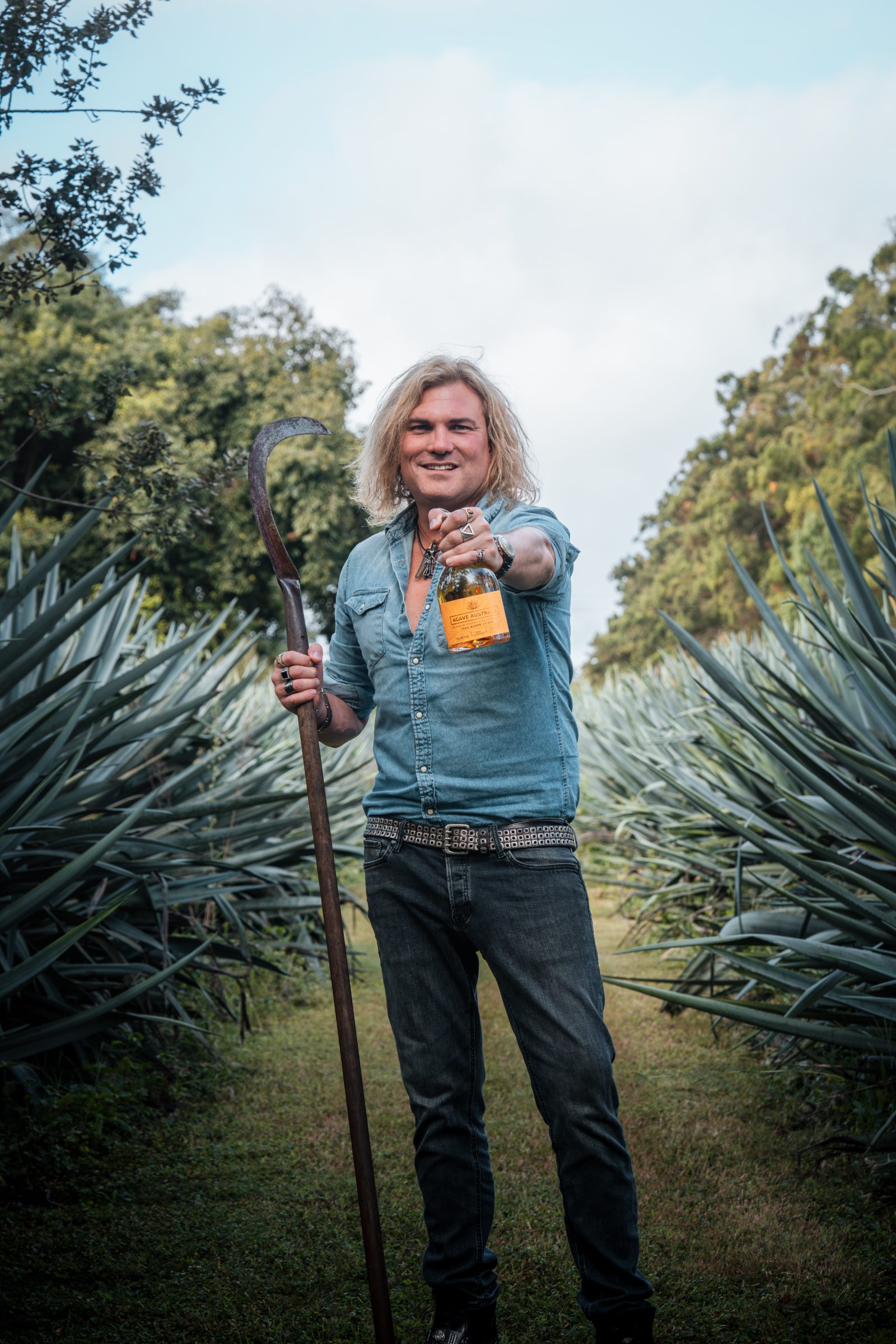 A man in the agave rows holding a long-handled coa upright, its curved blade clear against the sky
